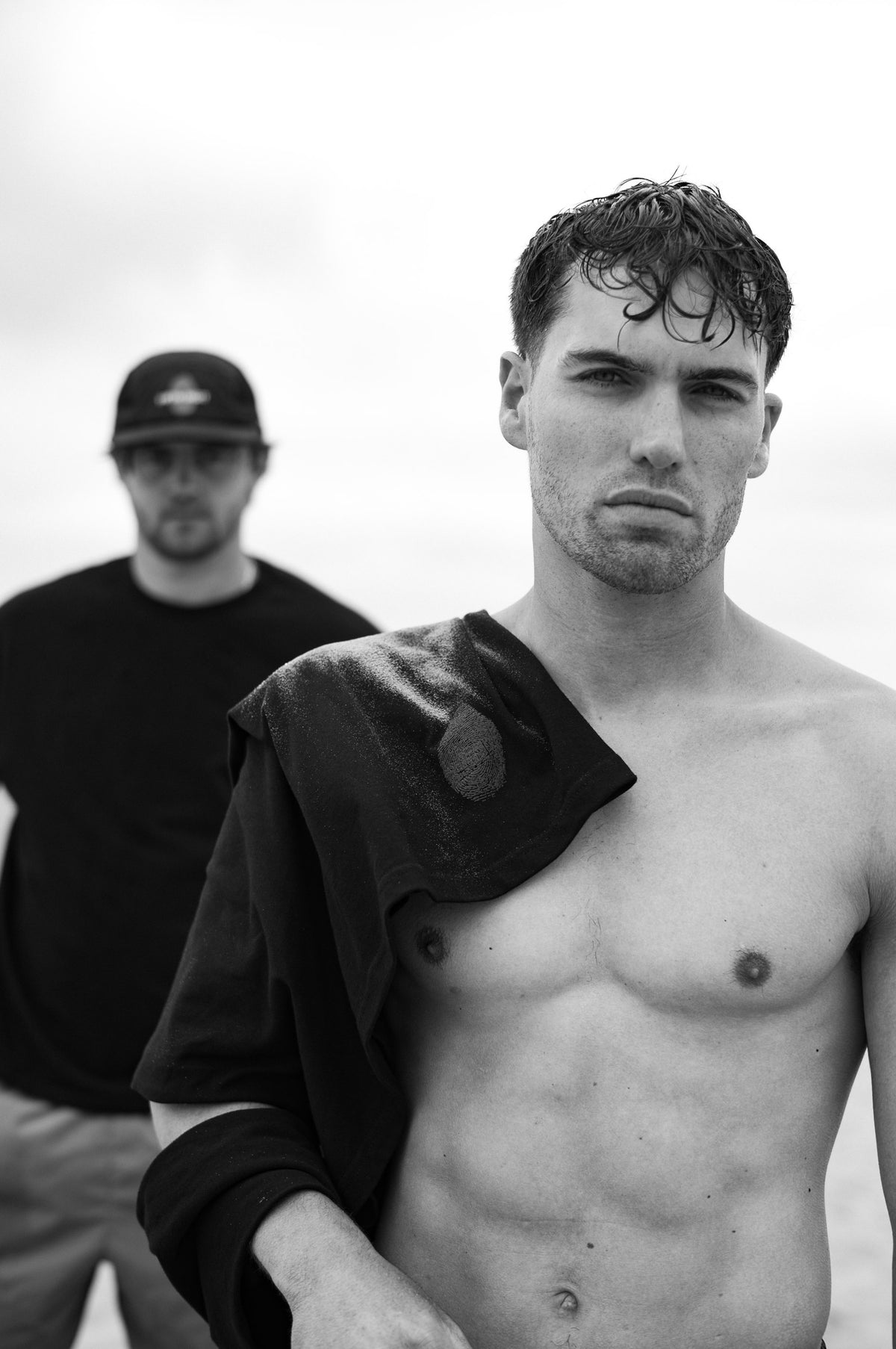 Two men on an overcast coastline. One in front, shirtless, a black tee over his shoulder. One behind in a black tee and cap. Black and white.