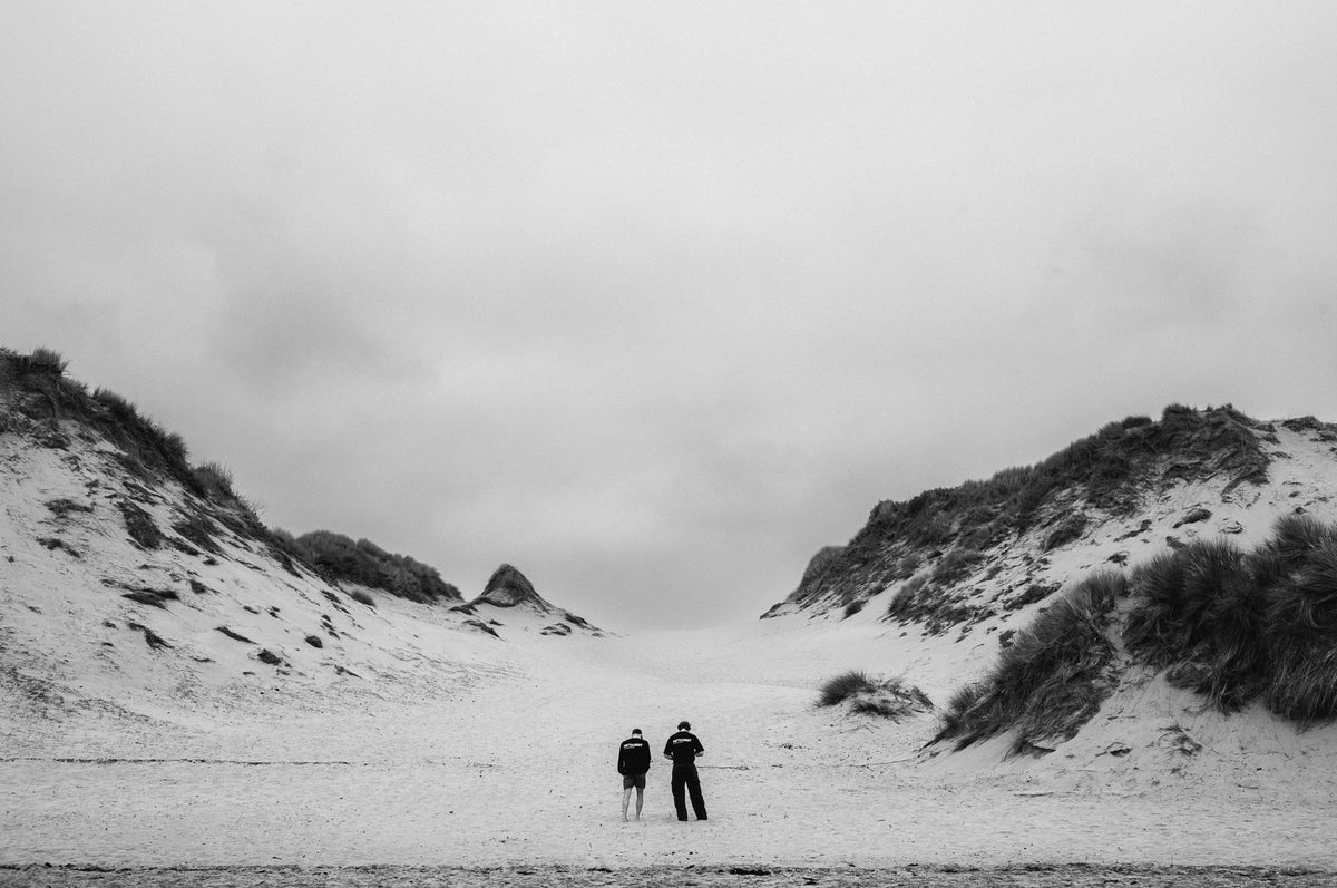 Two figures in black walk away between sand dunes under an overcast sky. Black and white.
