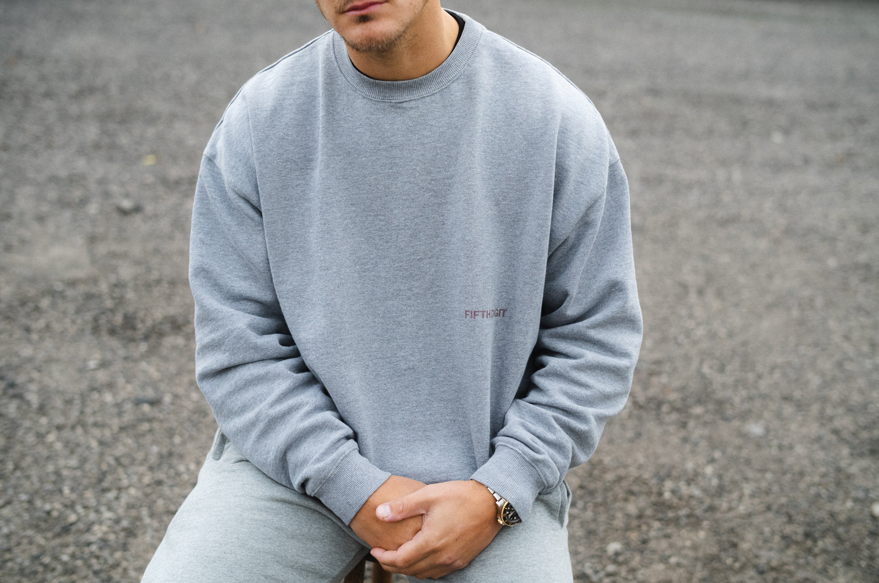 Model wearing Fifth Digit Arch 460gsm crewneck in grey marl, seated, front print detail