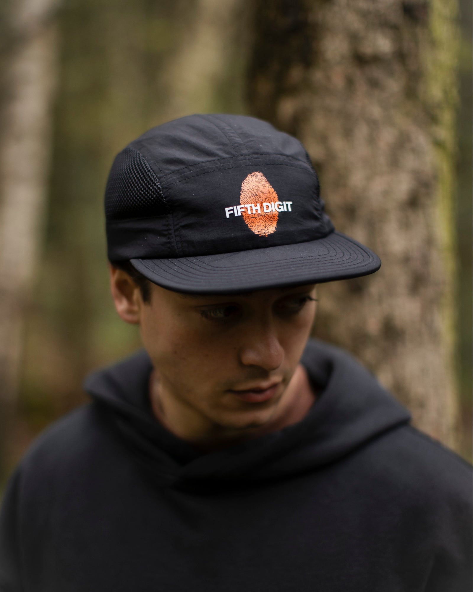 Fifth Digit Capsule 001 — black cap with orange thumbprint and Fifth Digit logo, woodland setting