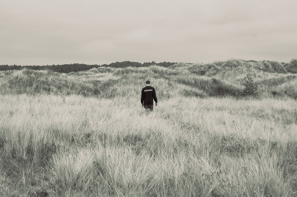 A lone figure in black walks into tall dune grass under an overcast sky.