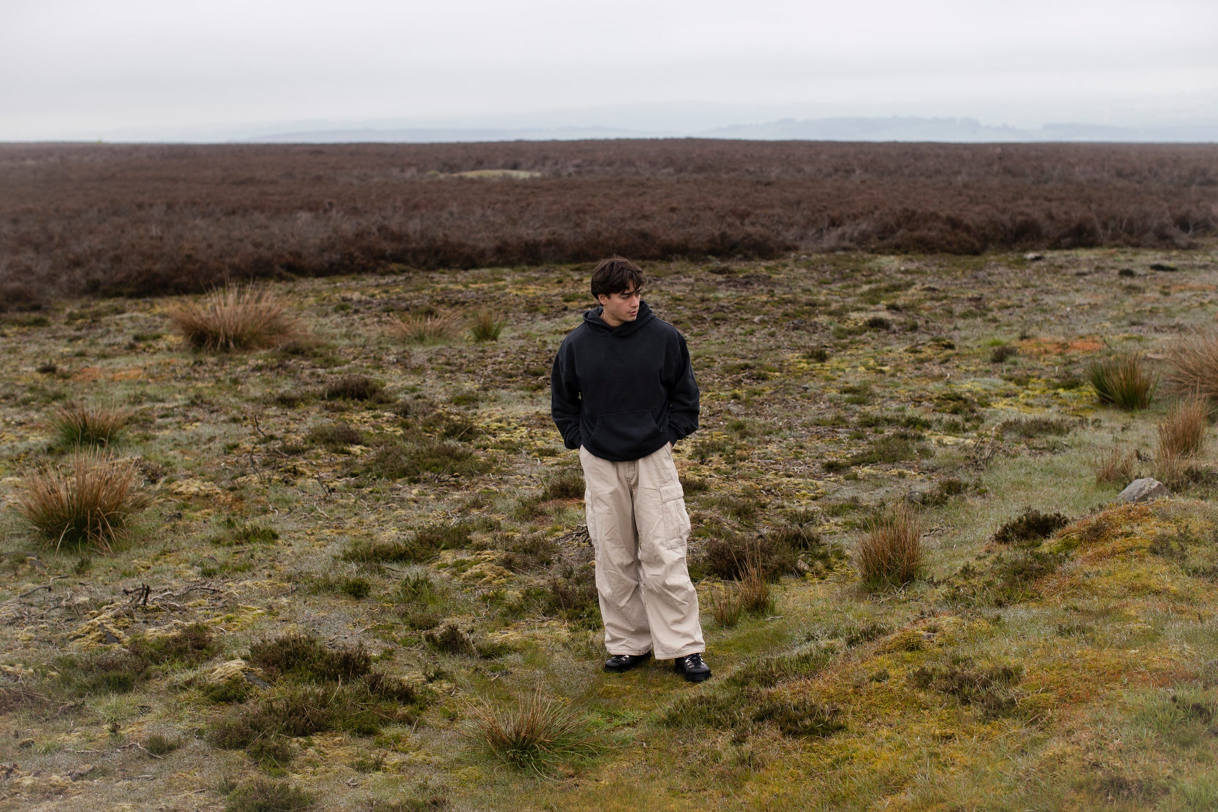 Fifth Digit Capsule 001 — figure in hoodie and cargo trousers, vast moorland, front view