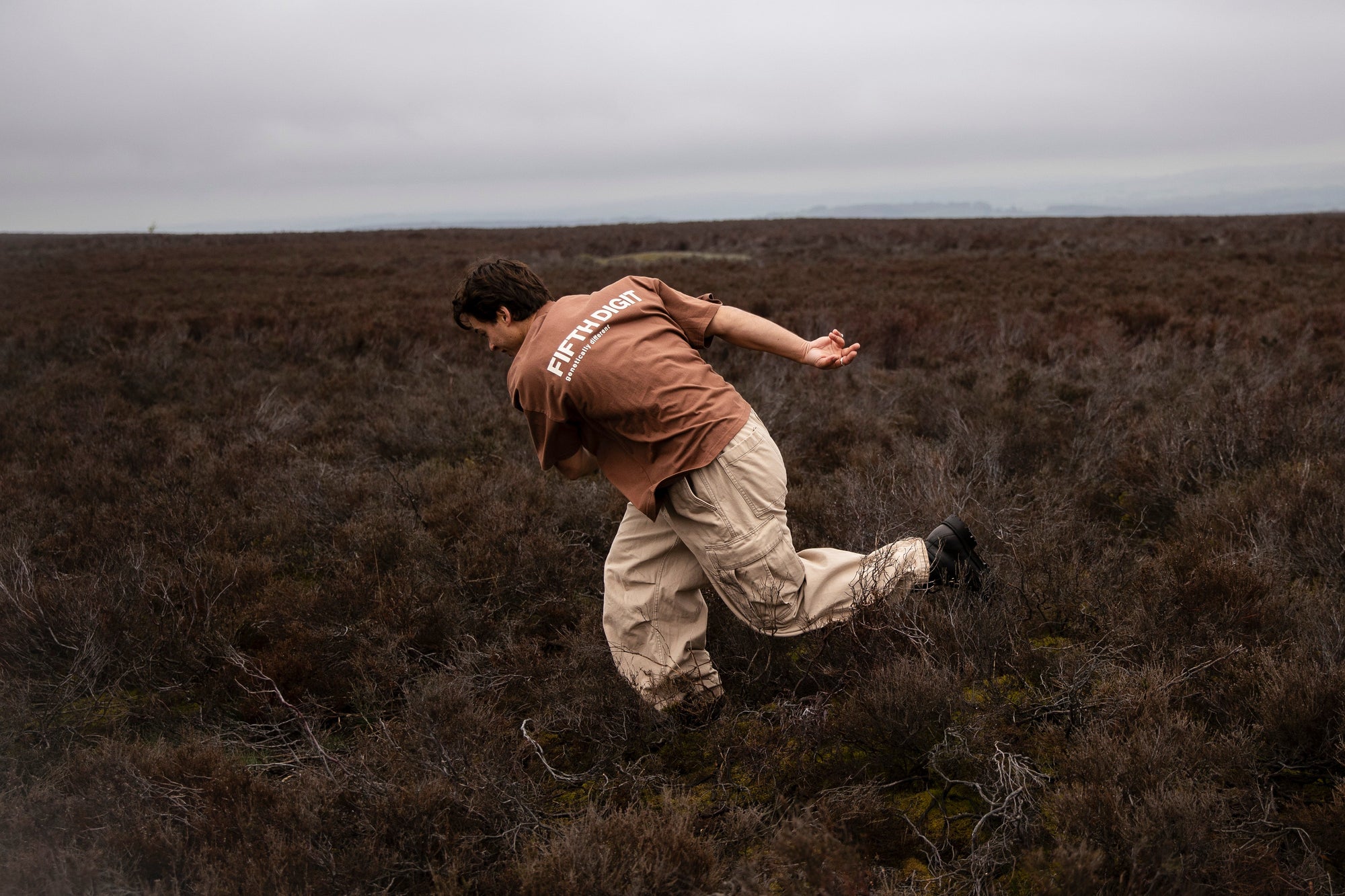 Fifth Digit Capsule 001 — figure in motion across heather moor, brown tee with back print