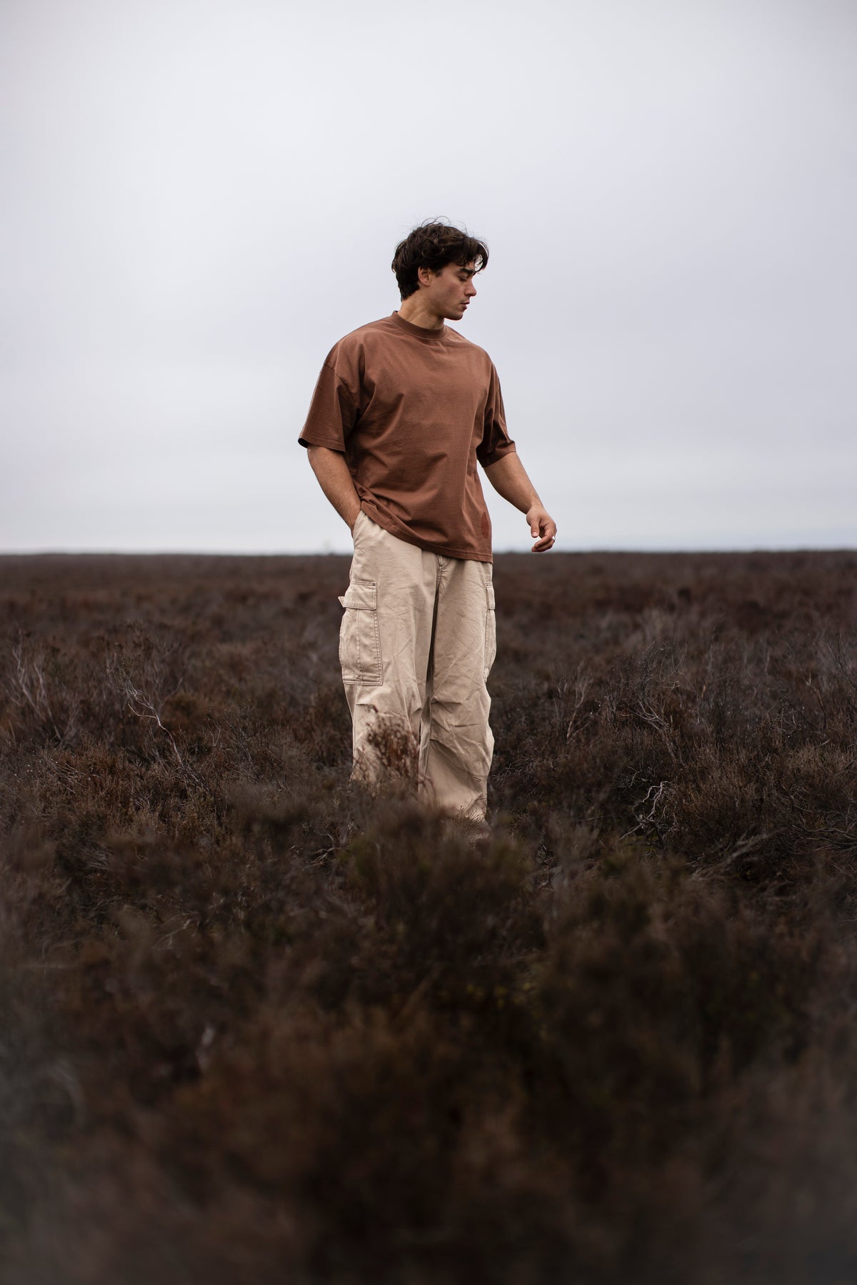 Fifth Digit Capsule 001 — figure in vintage brown tee and cream cargo trousers, moorland, side view