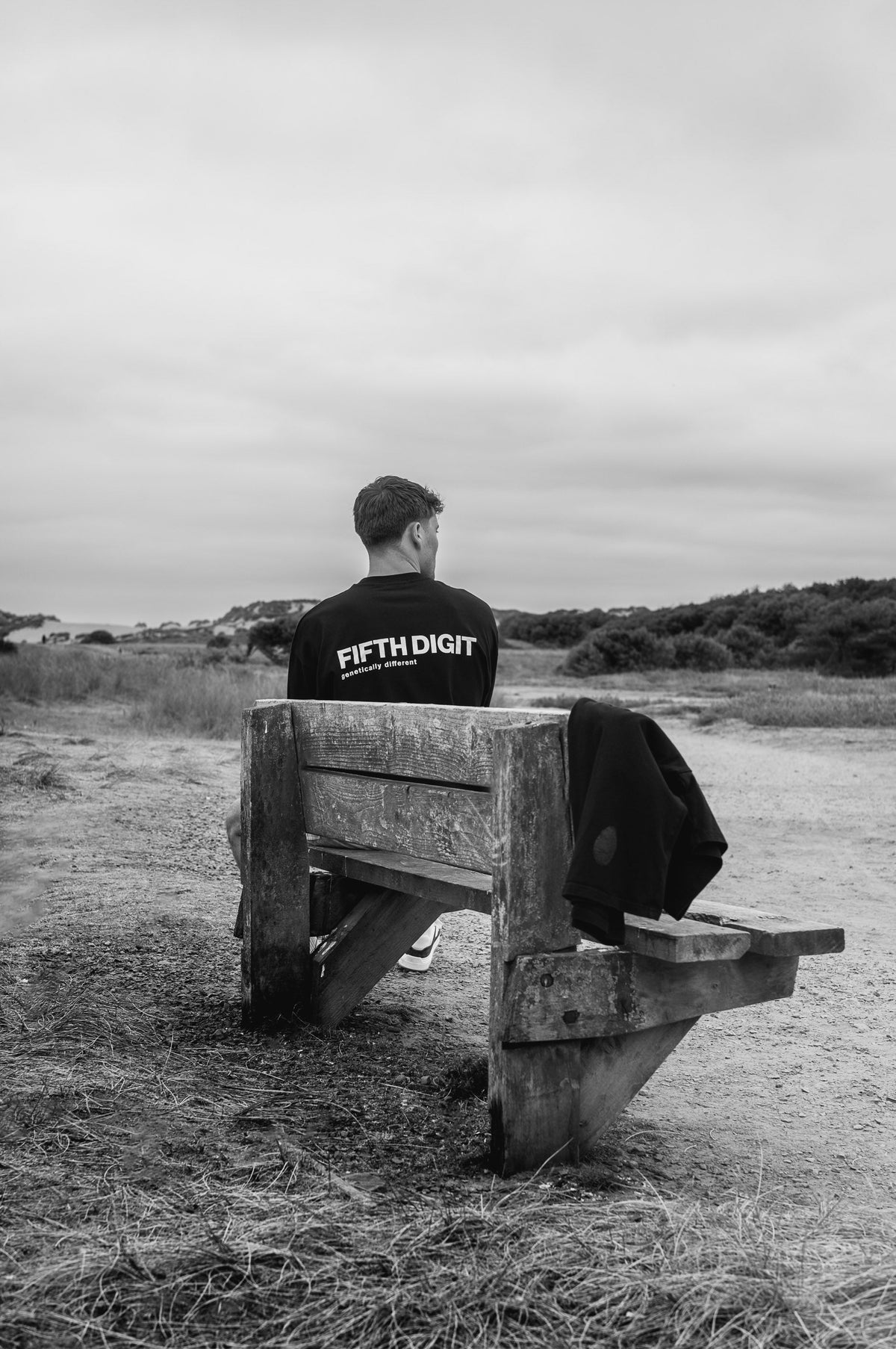 A man sits on a wooden bench in the dunes with his back turned, in a black Fifth Digit top with the back print. Black and white.