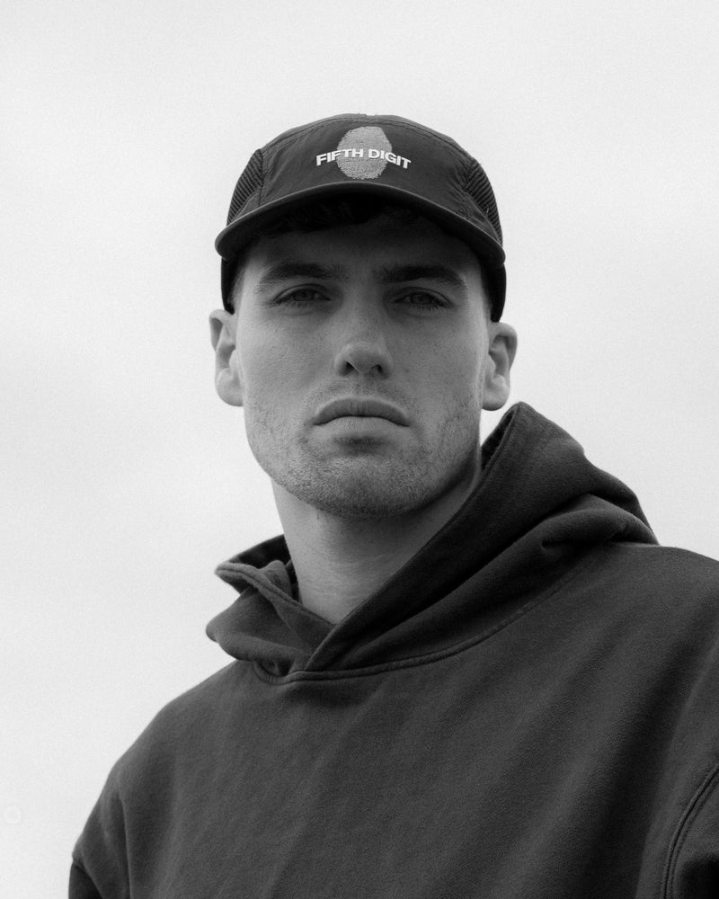 A man in a black Fifth Digit cap and a dark hoodie looks into the camera under an overcast sky. Black and white.