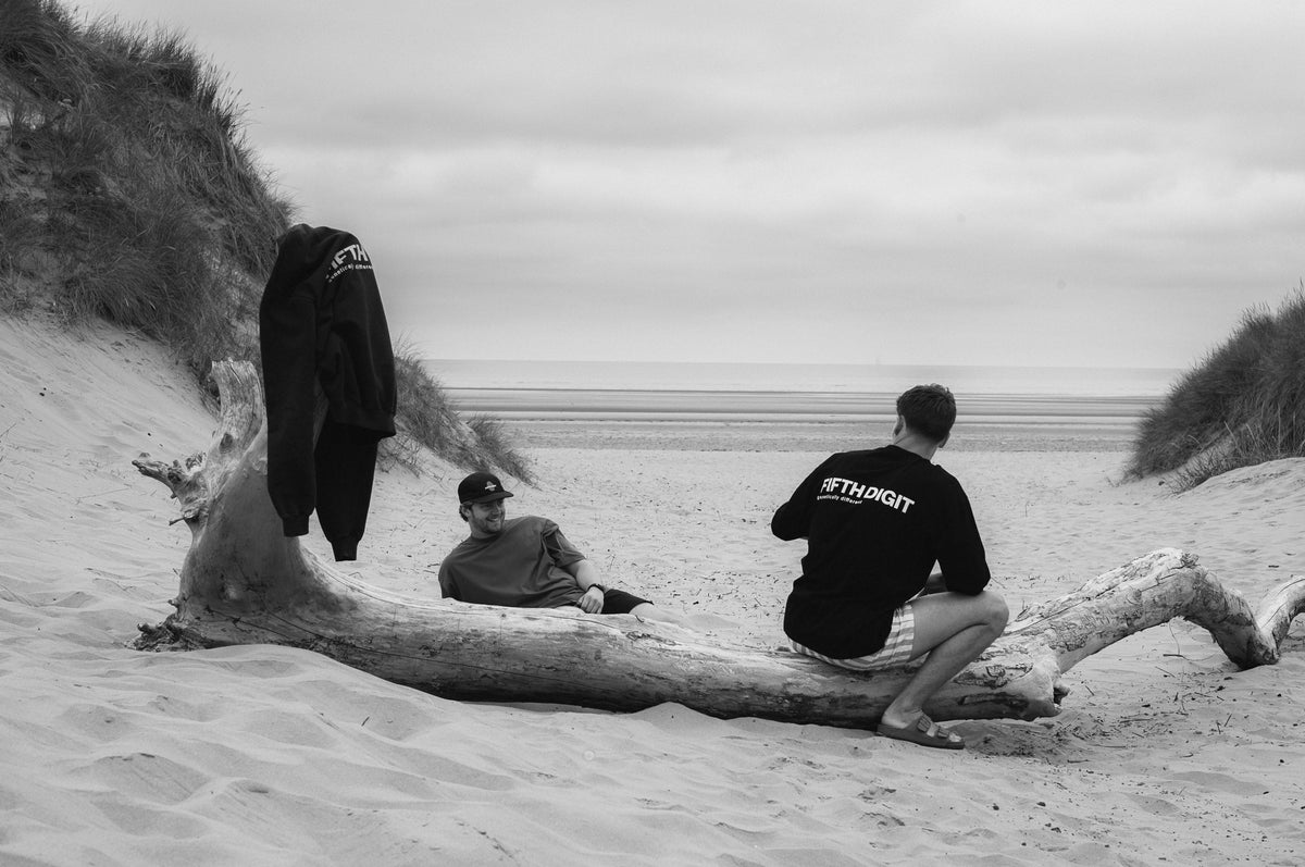 Two men on driftwood in the Formby dunes, the sea behind. One sits on the log in the Origin long sleeve, one leans back on the sand. A Frame hoodie hangs from a branch. Black and white.