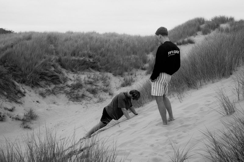 A man in a black Fifth Digit long sleeve watches his friend climb a steep dune on his hands. Black and white.