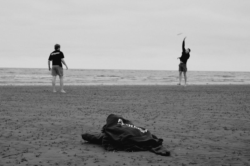 Two men throw a frisbee at the water's edge. A black Fifth Digit top lies on the sand in front of them. Black and white.
