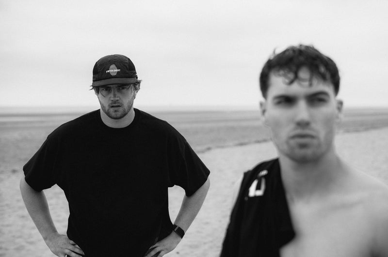 Two men on the beach, one in a black tee and cap with his hands on his hips, one shirtless beside him. Black and white.