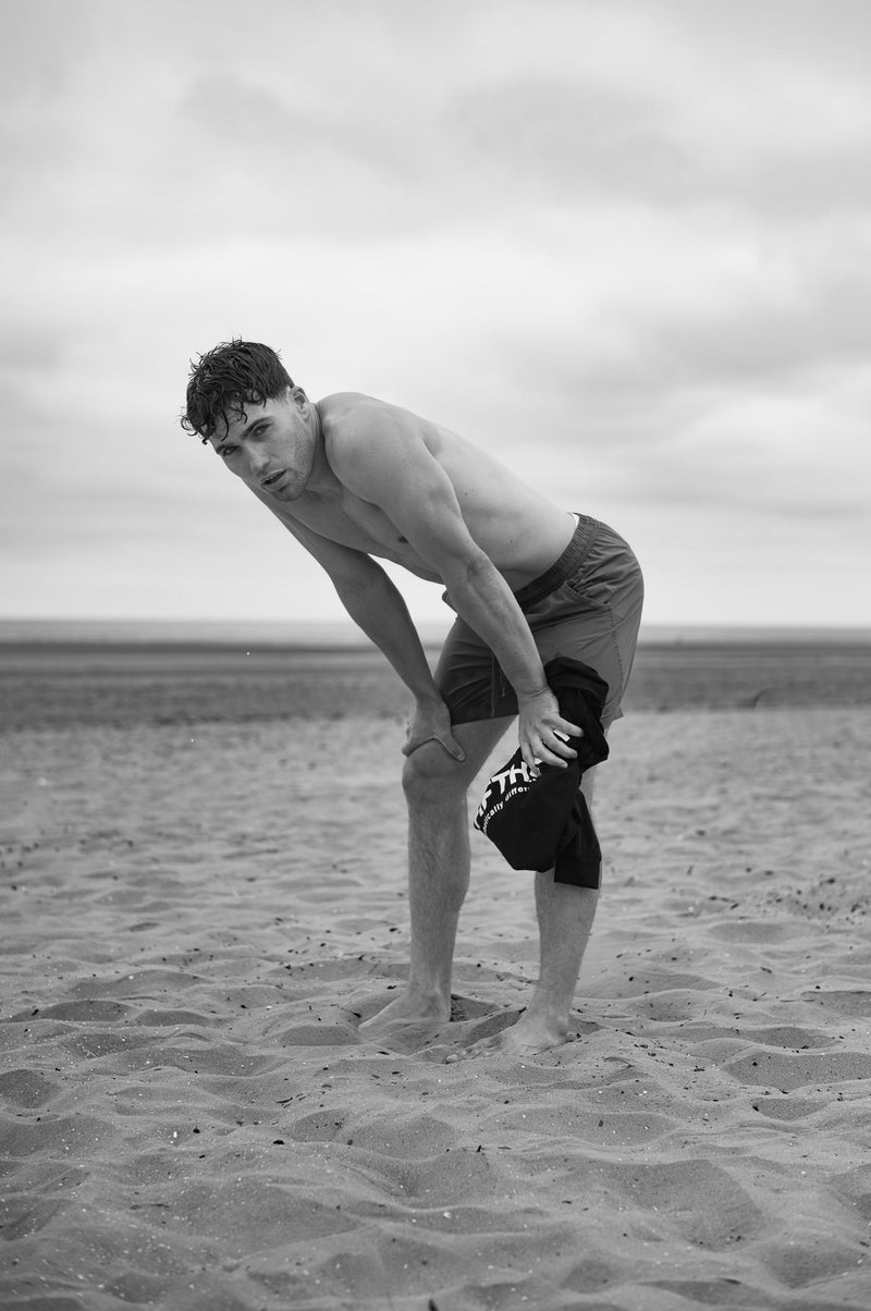 A shirtless man bends forward on the sand, hands on his knees, a black Fifth Digit top in his hand. Black and white.