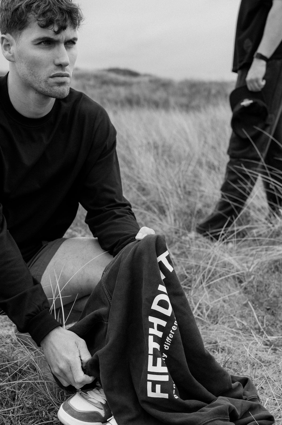 A man in a black long sleeve sits in the dune grass holding a black Fifth Digit hoodie, the back print showing. Black and white.