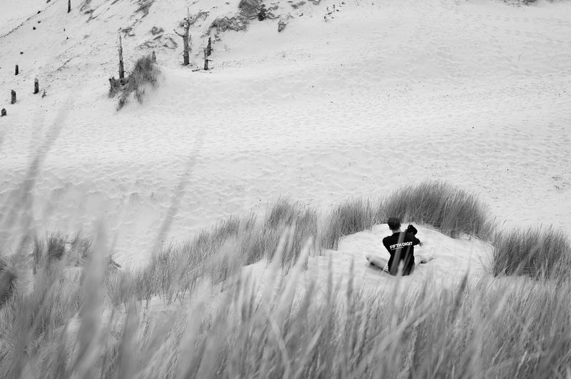 A man in a black Fifth Digit top sits in a hollow of the dunes, seen through tall grass. Black and white.