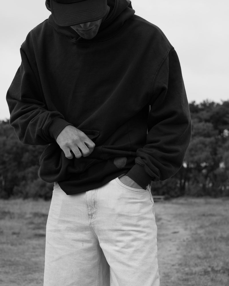 A man in a black Fifth Digit hoodie and light trousers adjusts the hem, standing in the grass. Black and white.