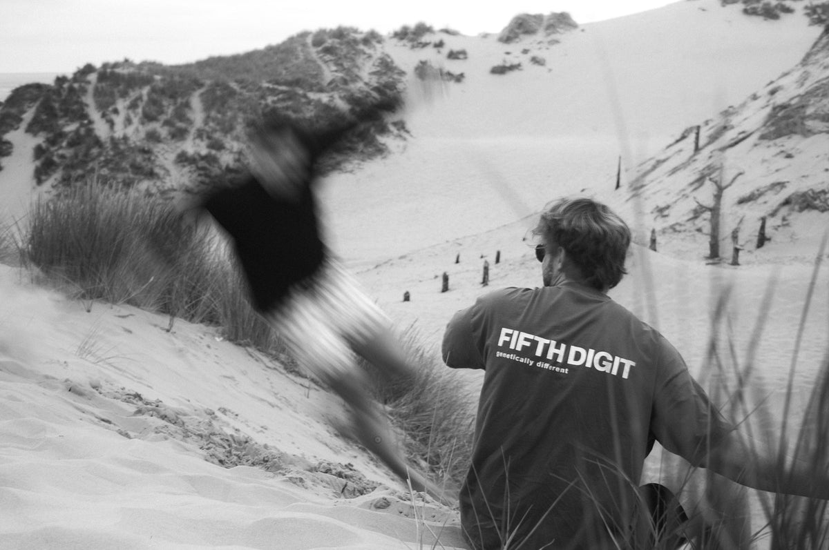 Two men in the dunes, one leaping past in a blur, the other in a Fifth Digit tee with the back print. Black and white.