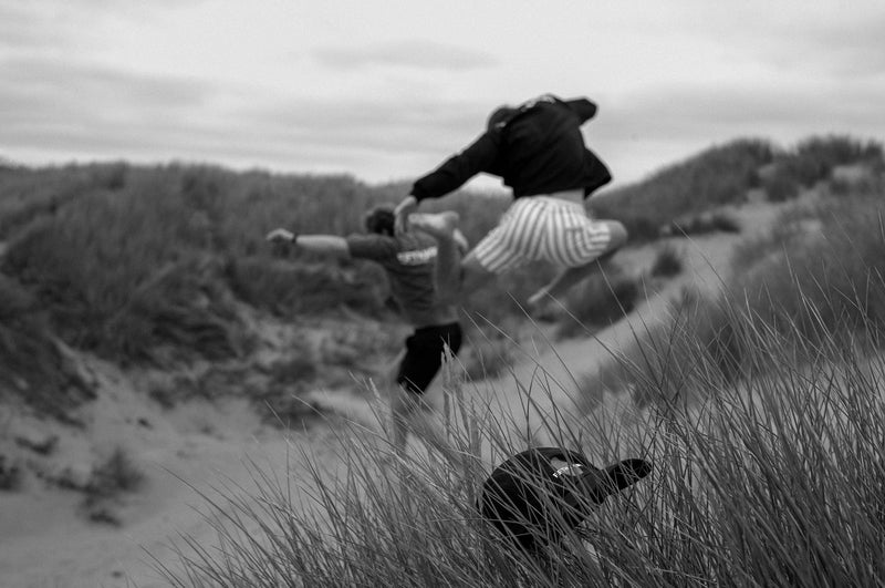 A man leaps over his friend in the dune grass, a black top lying on the sand below. Black and white.