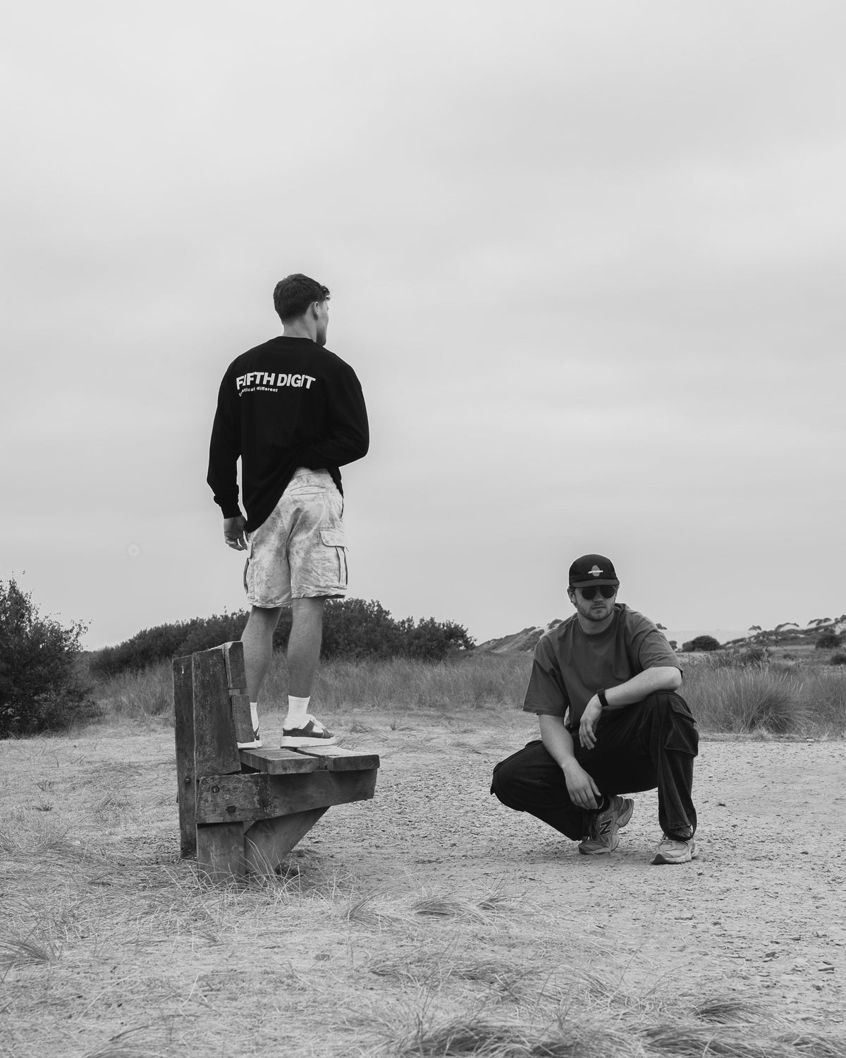 Two men on open dunes under an overcast sky. One stands on a wooden bench with his back turned, in the black Fifth Digit Origin long sleeve with the back print. One crouches beside him in the Pressure tee and Runner cap. Black and white.