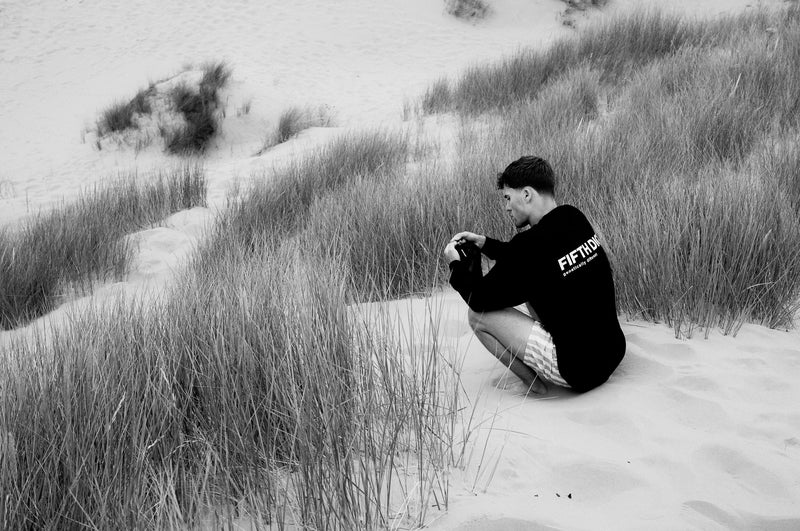 A man in a black Fifth Digit long sleeve crouches in the dune grass, the back print showing. Black and white.