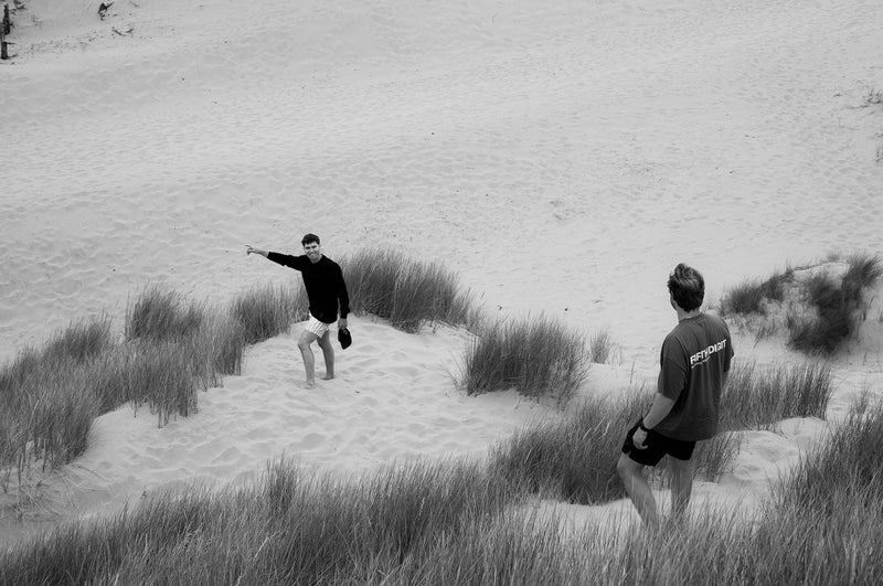 Two men in the dunes, one pointing out across the sand, the other watching from the grass. Black and white.