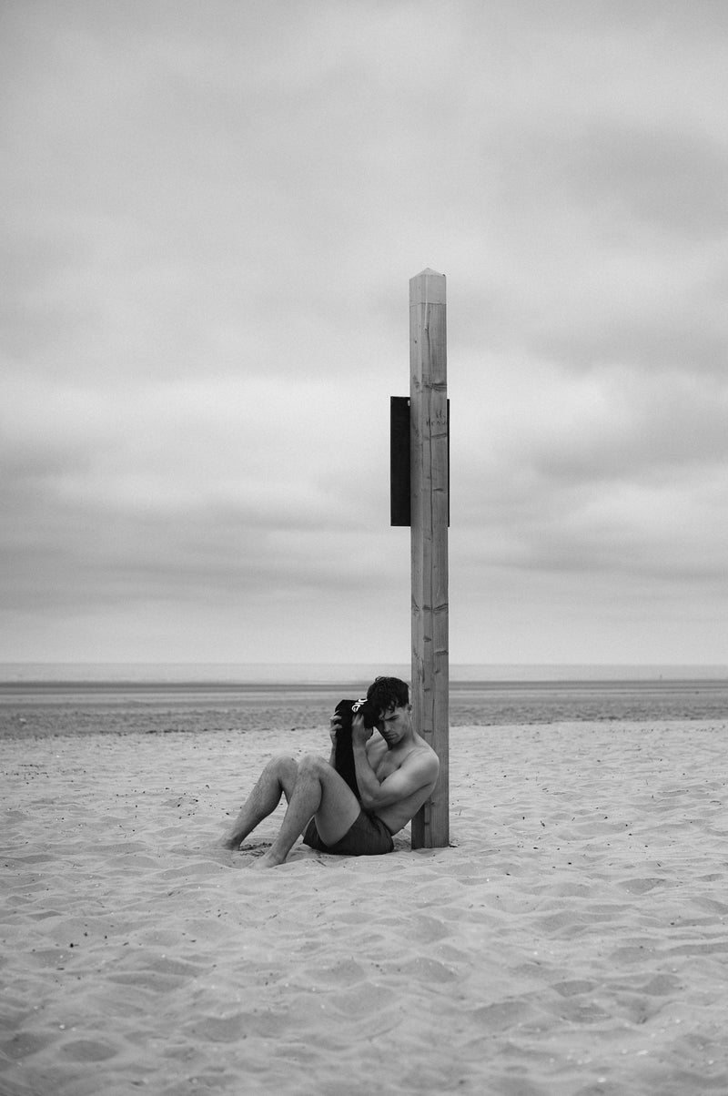 A man sits alone against a tall wooden post on an empty beach under an overcast sky. Black and white.