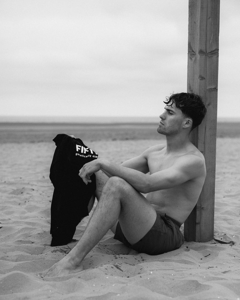 A shirtless man sits against a wooden post on the beach, a black Fifth Digit top hanging beside him. Black and white.