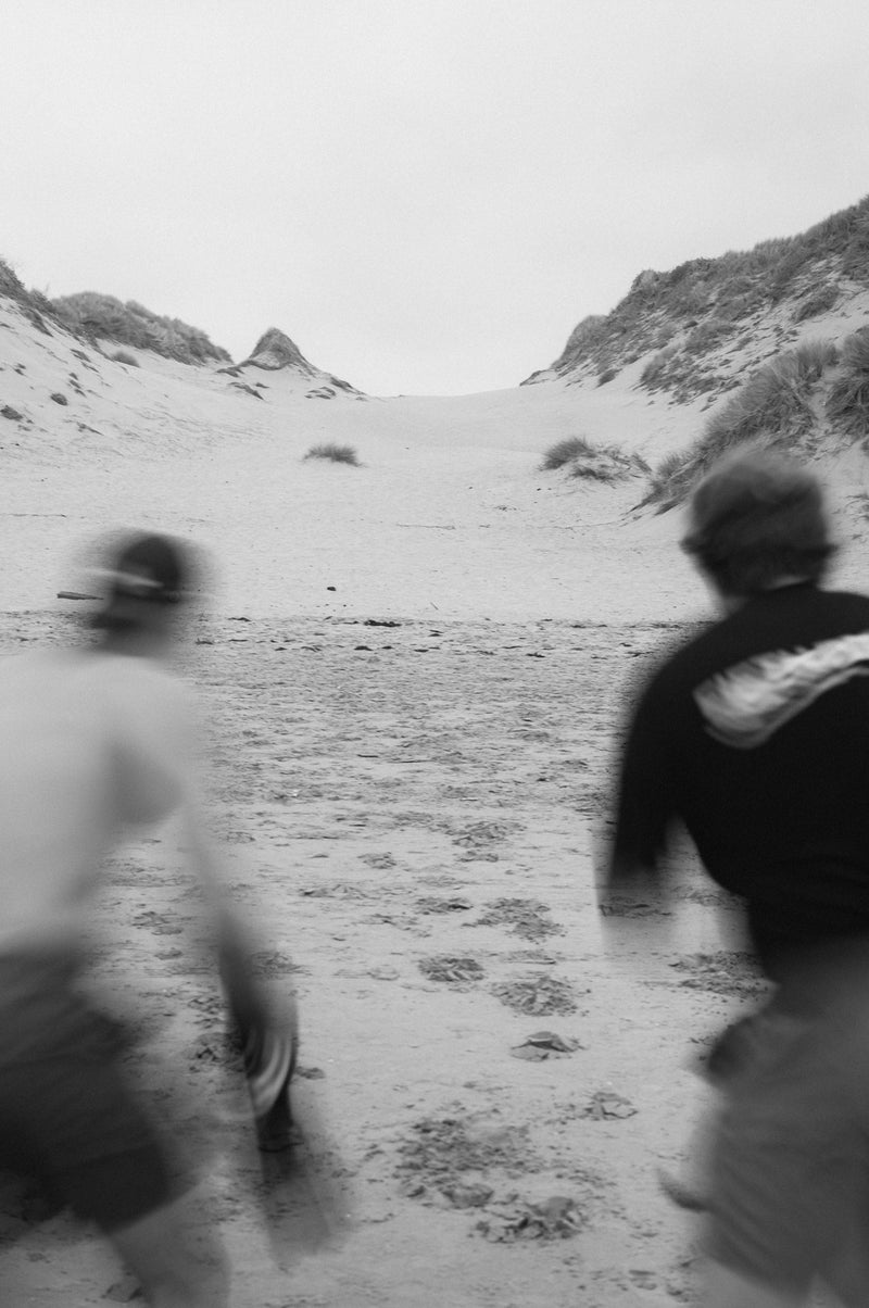Two men run across the sand towards the dunes, blurred by movement. Black and white.