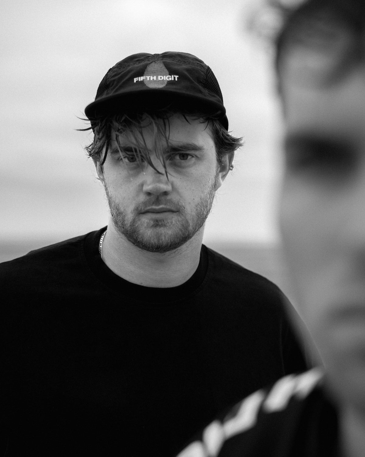 A man in the black Runner cap and a black tee looks into the camera, a friend blurred in the foreground. Black and white.