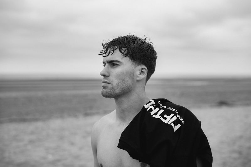 A shirtless man on the beach with a black Fifth Digit top over his shoulder, the print showing, looking out to sea. Black and white.