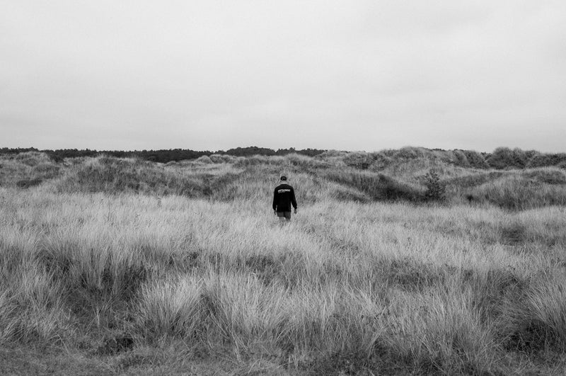 A lone figure in black walks into tall dune grass under an overcast sky. Black and white.