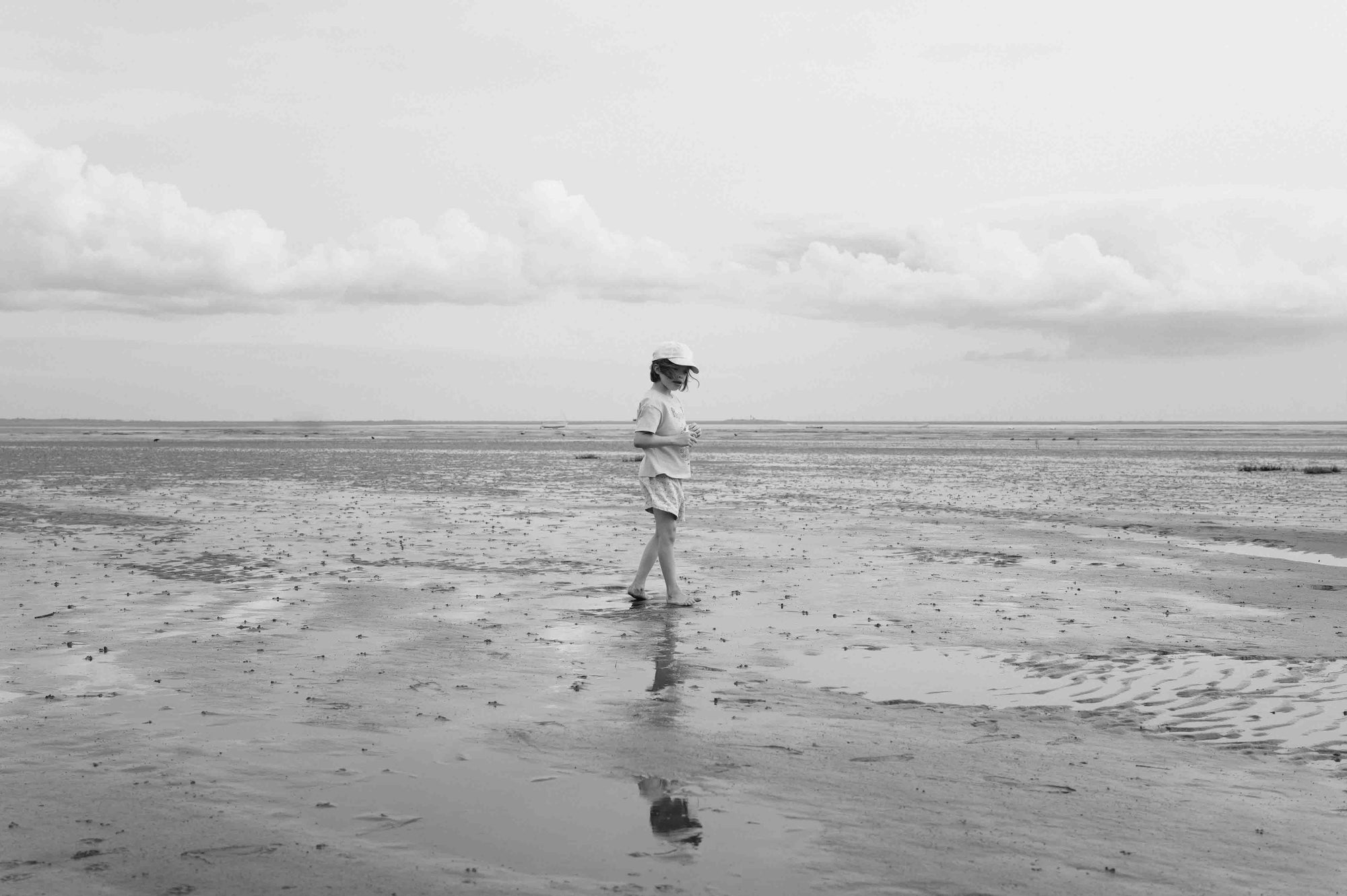 A small figure stands alone on a vast tidal flat. Sky and water meet at the horizon. Black and white.