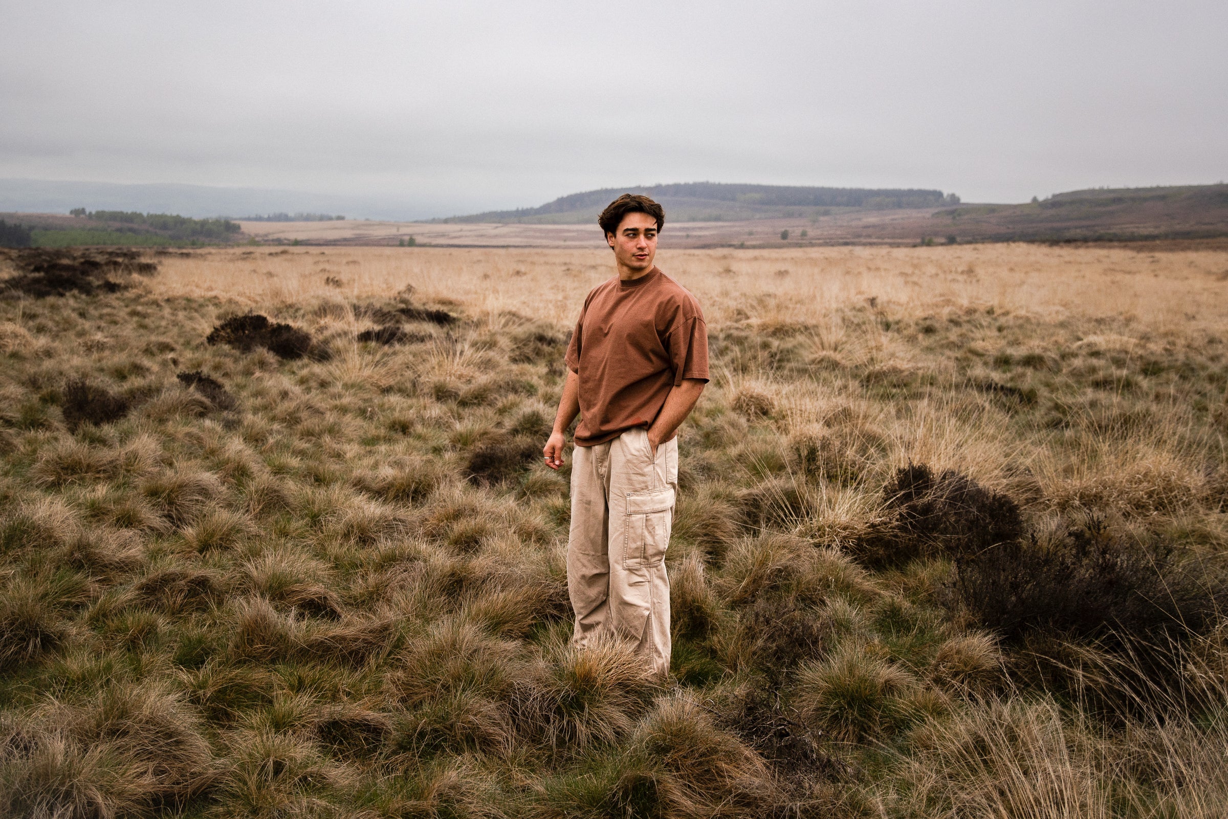 A man stands on open moorland wearing the Fifth Digit Pressure tee in vintage brown, wide landscape behind him, overcast sky.